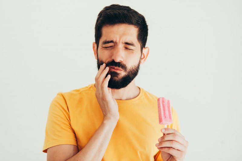 a man with a beard holding a popsicle while touching his cheek and wincing in pain because of a sensitive tooth