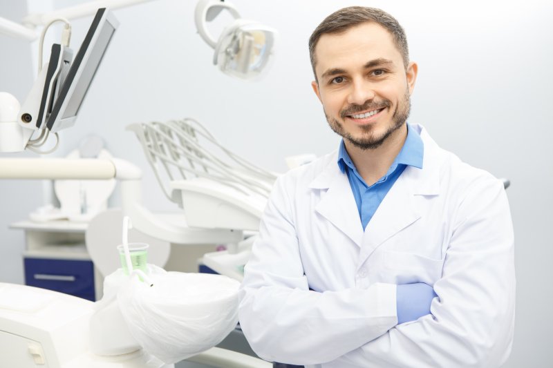 a male dentist wearing a lab coat and gloves while standing in a dental treatment room